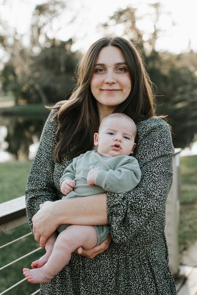 Kloe, a white woman with long brown hair, wearing a dress and holding a baby wearing a sweater.